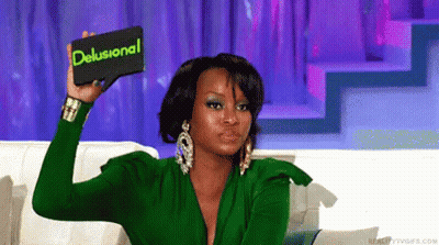 A Woman In A Green Dress Is Holding Up A Card That Says Delusional GIF
