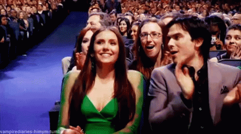 A Woman In A Green Dress Is Sitting Next To A Man In A Suit In Front Of A Crowd . GIF