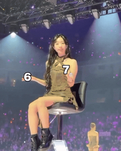A Woman In A Green Dress Is Sitting On A Chair With The Number 7 On Her Arm GIF