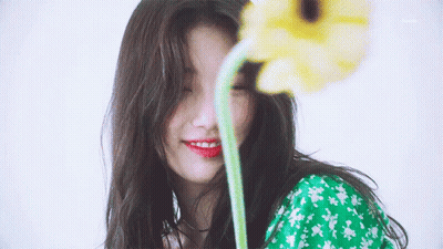A Woman In A Green Dress Is Smiling While Holding A Yellow Flower In Front Of Her Face GIF