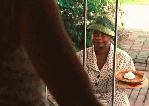 A Woman In A Green Hat Is Holding A Pie In Front Of A Window GIF