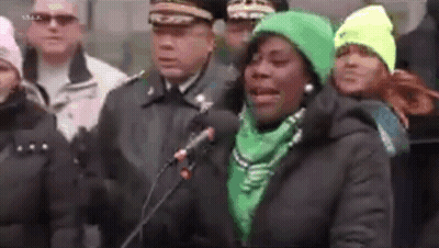 A Woman In A Green Hat Is Speaking Into A Microphone While Standing In Front Of A Crowd Of People . GIF