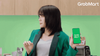 A Woman In A Green Jacket Is Holding A Cell Phone That Says Grab Mart GIF