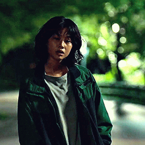 A Woman In A Green Jacket Is Standing On A Street . GIF