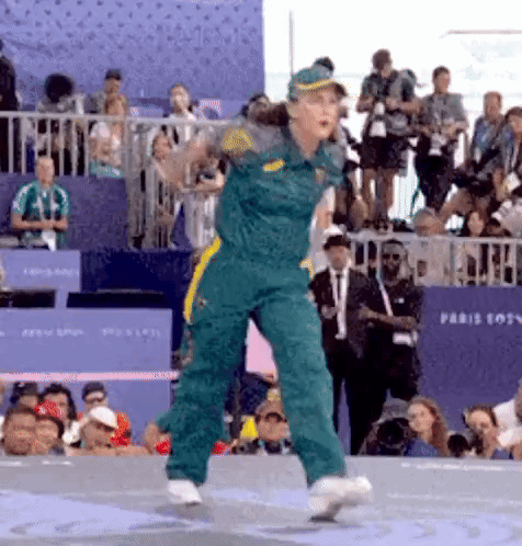A Woman In A Green Jumpsuit Is Dancing In Front Of A Crowd GIF
