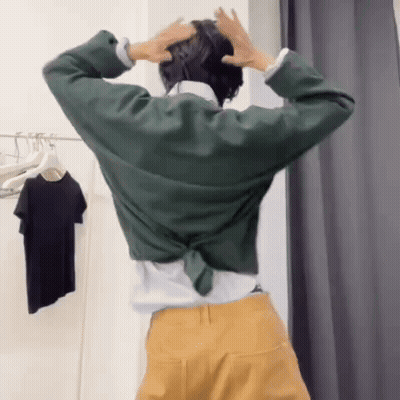 A Woman In A Green Shirt And Yellow Pants Is Standing In A Dressing Room With Her Hands On Her Head GIF