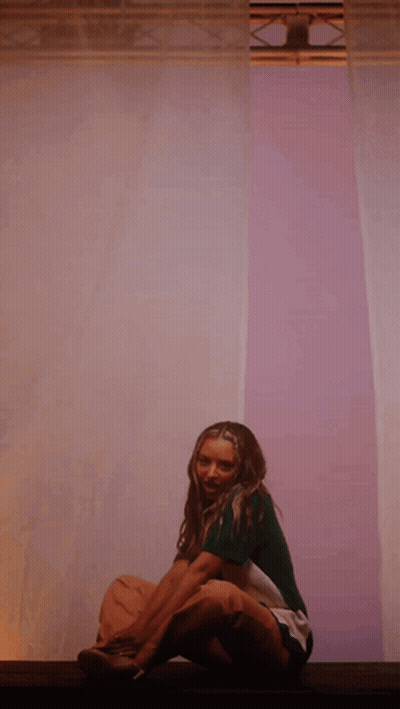 A Woman In A Green Shirt Sits On A Purple Wall GIF