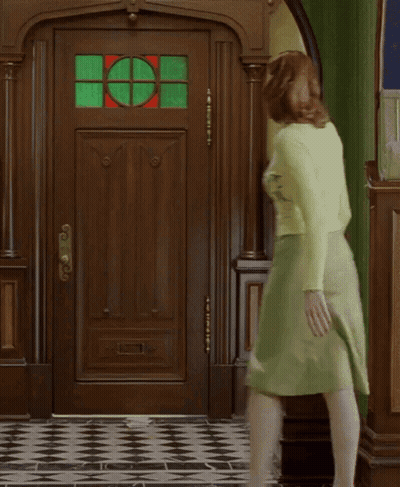 A Woman In A Green Skirt Is Walking In Front Of A Door With A Stained Glass Window GIF