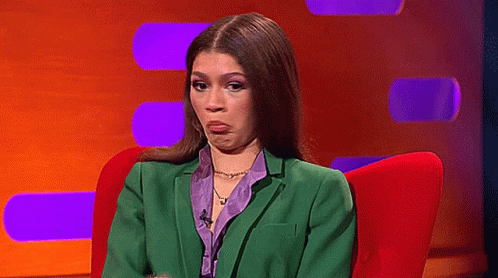 A Woman In A Green Suit And Purple Shirt Is Sitting In A Red Chair And Making A Funny Face . GIF