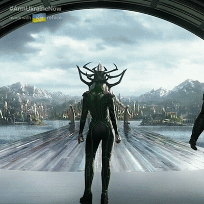 A Woman In A Green Suit Is Standing In Front Of A Body Of Water And Mountains GIF
