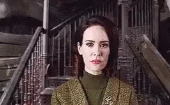 A Woman In A Green Suit Is Standing In Front Of A Staircase . GIF