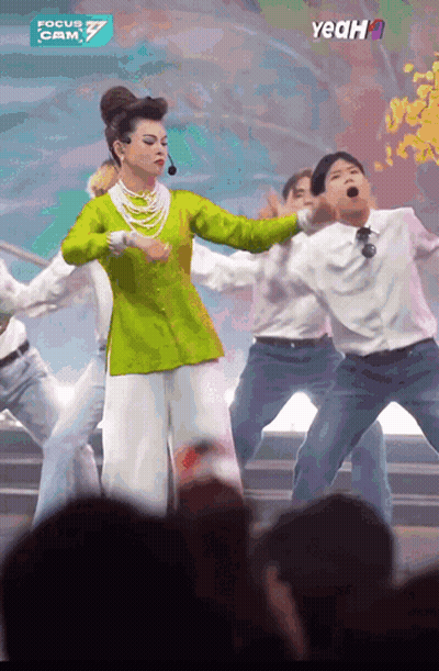 A Woman In A Green Top And White Pants Is Dancing In Front Of A Crowd And A Sign That Says Yeah GIF