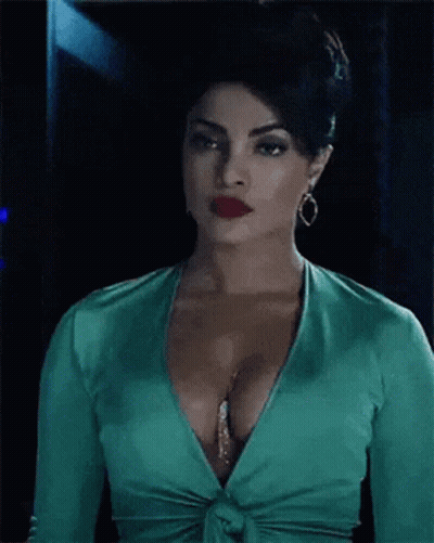 A Woman In A Green Top With A Plunging Neckline And Red Lips Is Standing In A Dark Room . GIF
