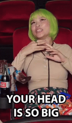 A Woman In A Green Wig Is Sitting In A Movie Theater Eating Chips And Drinking A Drink . GIF