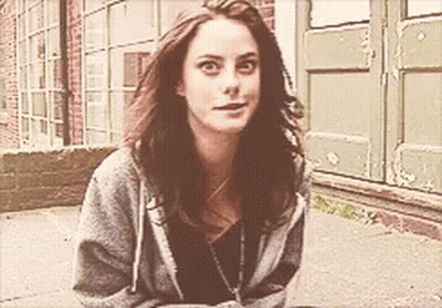 A Woman In A Grey Hoodie Is Sitting On The Sidewalk In Front Of A Building GIF