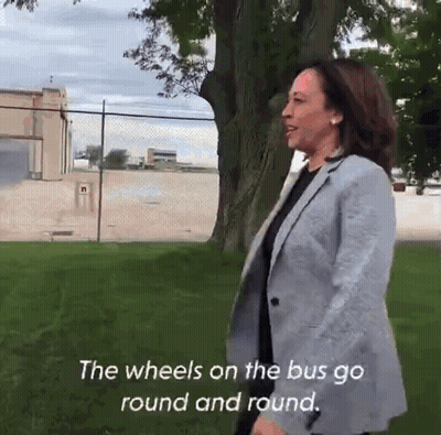 A Woman In A Grey Jacket Is Standing In A Grassy Field With The Words " The Wheels On The Bus Go Round And Round " Below GIF