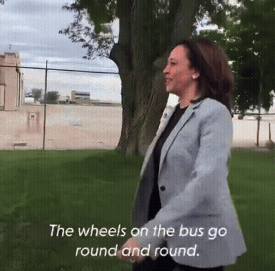 A Woman In A Grey Jacket Is Walking In A Park With The Words " The Wheels On The Bus Go Round And Round " Above Her GIF