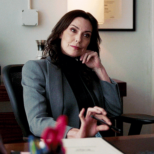 A Woman In A Grey Jacket Sits At A Desk With A Pen In Her Hand GIF