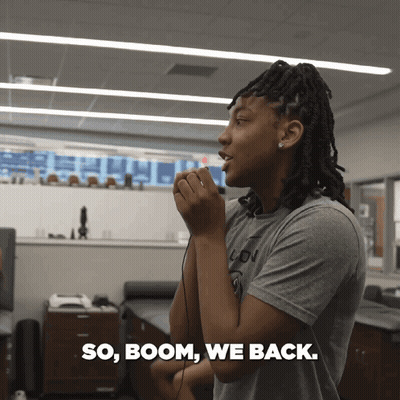 A Woman In A Grey Shirt Is Talking Into A Microphone With The Words So Boom We Back Written Below Her GIF