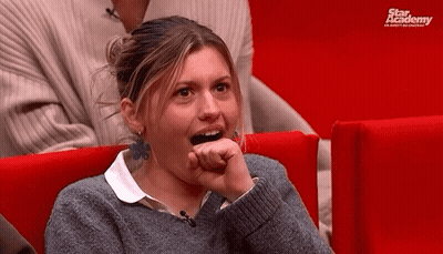 A Woman In A Grey Sweater Is Sitting In A Red Chair With A Star Academy Logo Behind Her GIF