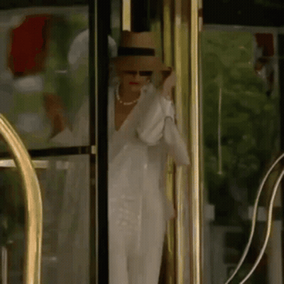 A Woman In A Hat And Sunglasses Is Standing In Front Of A Glass Door . GIF