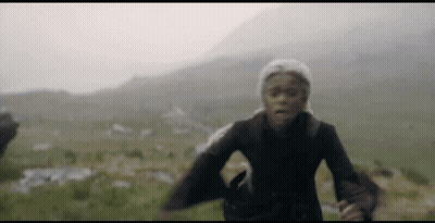 A Woman In A Hat Is Running In A Field With Mountains In The Background . GIF