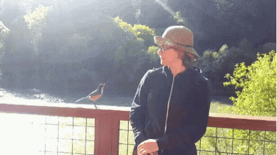 A Woman In A Hat Stands Next To A Bird On A Railing Overlooking A River GIF