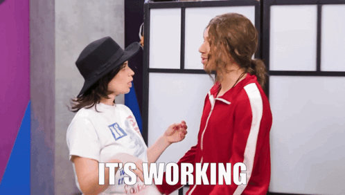 A Woman In A Hat Talks To A Man In A Red Jacket With The Words " It 'S Working " On The Bottom GIF