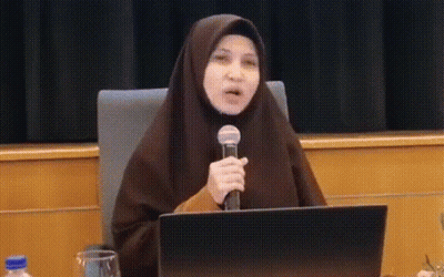 A Woman In A Hijab Is Sitting At A Table Holding A Microphone And A Laptop . GIF