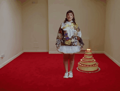 A Woman In A Hoodie Stands In Front Of A Cake With Candles On It GIF