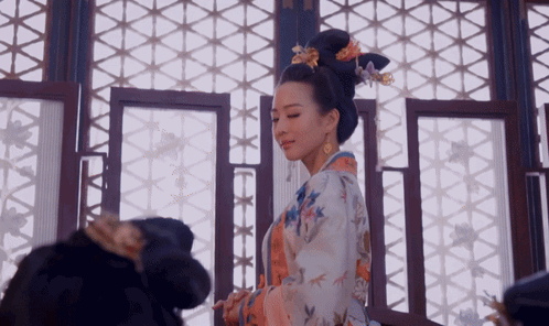 A Woman In A Kimono With Flowers In Her Hair Is Standing In Front Of A Window GIF