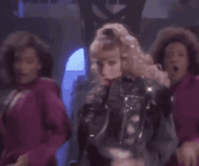 A Woman In A Leather Jacket Is Dancing In Front Of A Group Of Dancers In Pink Dresses . GIF