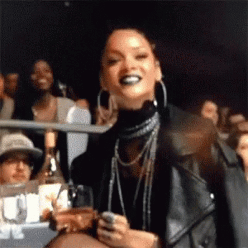 A Woman In A Leather Jacket Is Standing In A Crowd Holding A Glass Of Wine And A Cell Phone . GIF