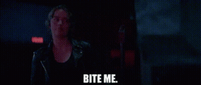 A Woman In A Leather Jacket Is Standing In A Dark Room With The Words Bite Me Written On The Screen . GIF