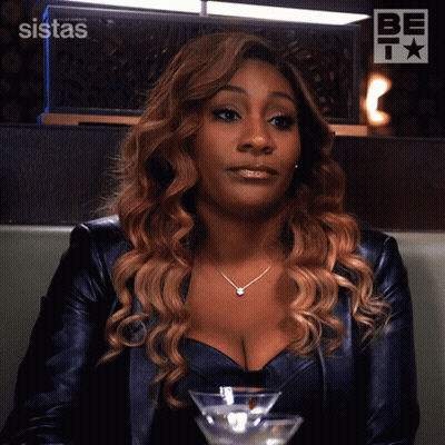 A Woman In A Leather Jacket Sits At A Table With A Martini In Front Of Her And A Sign That Says Sistas GIF