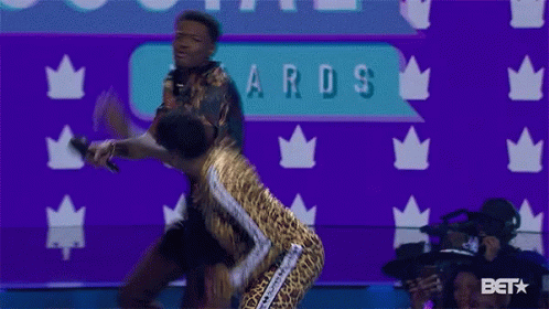 A Woman In A Leopard Print Outfit Stands Next To A Man Holding A Microphone At An Awards Show GIF