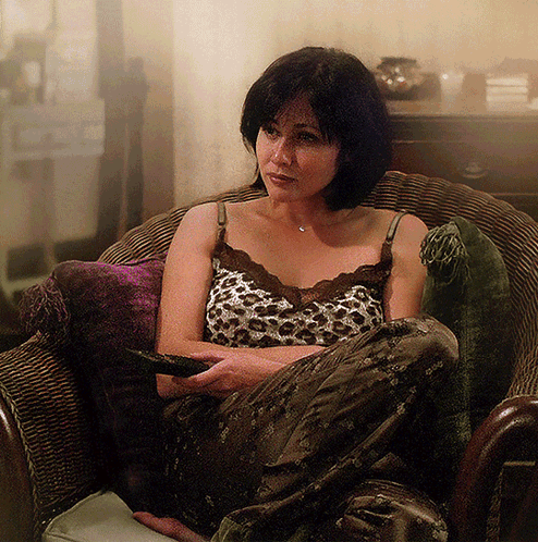 A Woman In A Leopard Print Tank Top Sits In A Chair Holding A Remote Control GIF