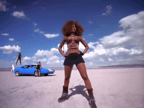 A Woman In A Leopard Print Top And Shorts Stands In The Sand Near A Blue Car GIF