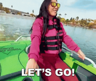 A Woman In A Life Jacket And Sunglasses Is Sitting In A Boat On A Lake And Says Let 'S Go . GIF