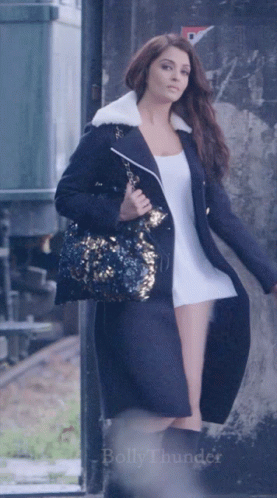 A Woman In A Long Black Coat Is Holding A Purse And The Word Bollythunder Is On The Bottom Of The Image GIF