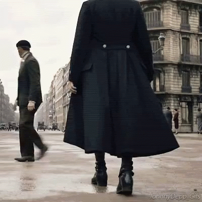 A Woman In A Long Black Coat Is Walking Down A Street While A Man In A Suit Walks Behind Her . GIF