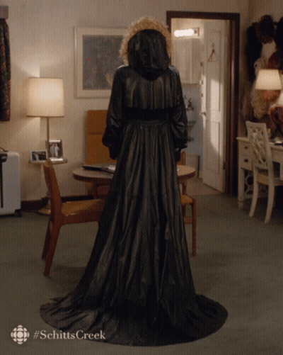 A Woman In A Long Black Coat With The Word Schitts Creek On The Bottom Right GIF