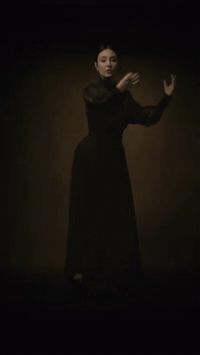 A Woman In A Long Black Dress Is Standing In The Dark With Her Hands Outstretched . GIF