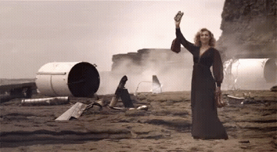 A Woman In A Long Black Dress Is Standing On A Rocky Beach Holding A Bottle In Her Hand GIF