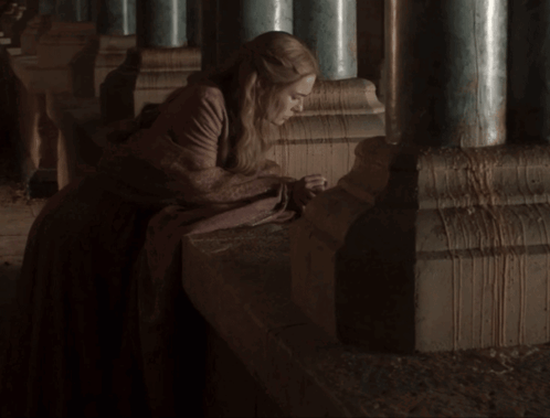A Woman In A Long Brown Dress Sits On A Ledge Between Two Columns GIF