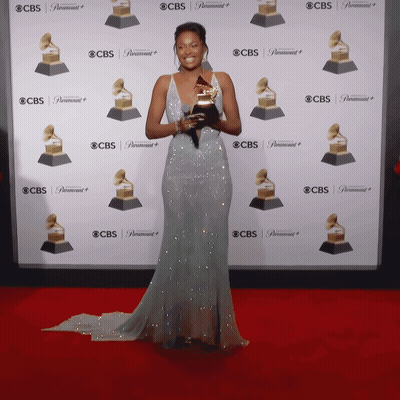 A Woman In A Long Dress Holds A Grammy Trophy In Front Of A Wall That Says Cbs GIF