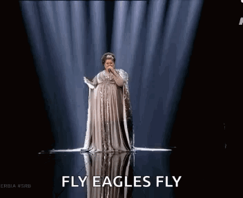 A Woman In A Long Dress Is Singing Into A Microphone On A Stage With The Words Fly Eagles Fly Above Her GIF