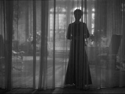 A Woman In A Long Dress Is Standing Behind A Curtain In A Black And White Photo GIF