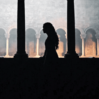 A Woman In A Long Dress Is Standing In Front Of A Window In A Dark Room . GIF