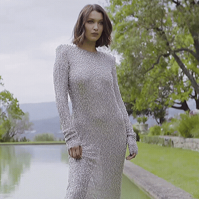 A Woman In A Long Silver Dress Is Standing In Front Of A Body Of Water GIF